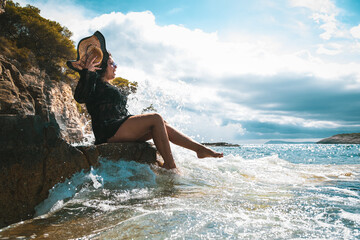 Brunette girl sitting on a rock by the sea on a sunny summer day in Croatia, suprised and shocked by the wave hitting the shore and splashing her. Fun on the beach while traveling in 2021