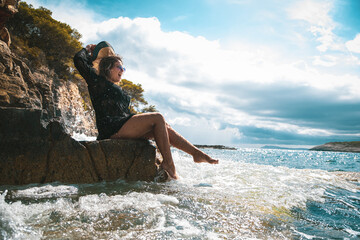 Brunette girl sitting on a rock by the sea on a sunny summer day in Croatia, suprised and shocked by the wave hitting the shore and splashing her. Fun on the beach while traveling in 2021