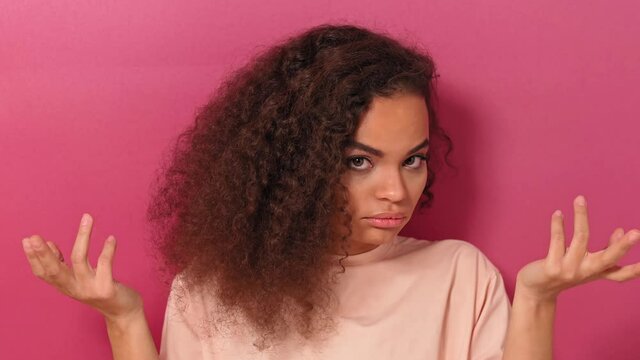 Confused, i dont know gesture charming nodding her head African American woman with long curly hair looking at camera wearing peachy t-shirt isolated on pink background. 4K footage. 