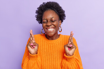 Positive dark skinned curly young woman smiles broadly makes wish prays with good attitude waits...