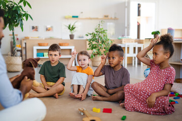 Group of small nursery school children with man teacher sitting on floor indoors in classroom, raising hands.