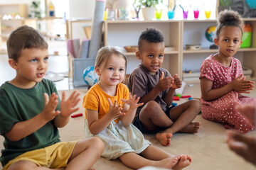 Group of small nursery school children sitting on floor indoors in classroom, clapping. - obrazy, fototapety, plakaty