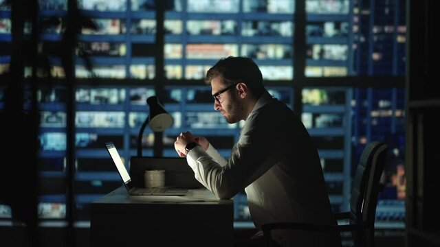 Serious man employee sit at desk develop start up ideas, search problem solution working hard use laptop app at night in modern skyscrapers office room. Deadline, careerism, workaholism concept