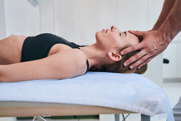 Fototapeta na wymiar Young elegant Caucasian woman in black sports bra enjoying facial massage in medical office