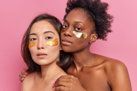 Two young multiethnic women apply golden patches under eyes stand closely to each other have healthy clean soft skin enjoy spa and beauty day isolated on pink wall. Face care treatment at home - Powered by Adobe