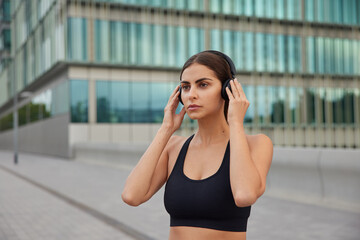 Serious determined brunette European woman in sporty black top listens music via wireless headphones focused into distance goes in for sport poses in urban setting going to have workout outdoors.
