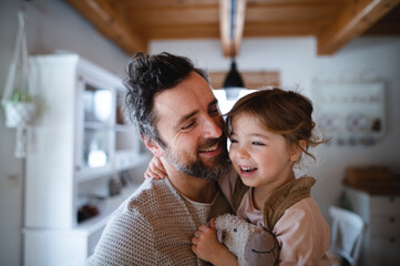 Mature father with small daughter standing indoors at home, holding and hugging.