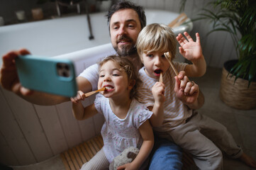 Father with two small children brushing teeth indoors at home, taking selfie.