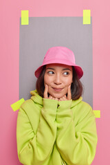 Vertical image of thoughtful Asian girl forces smile keeps index figers near corners of lips thinks about something nice wears pink panama green sweatshirt poses indoor. Human face expressions