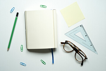 notebook with space for text, desk, reminder note, yellow note, blue bookmark, brown glasses, green pencil, ruler, paperclips, on a white background, photo from above