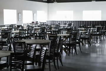 Empty ballroom, with several round tables, no decorations, and dark brown chairs. In the background the windows with the light almost bursting.