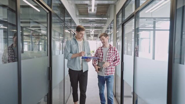 Male designers discussing work, walking in office corridor, advertising agency
