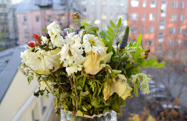 A wilted frozen bouquet of white roses and chrysanthemums, greenery on open window against the background of the street and houses. The end of holiday, relations