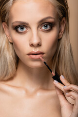 sensual woman with green eyes applying pink lip gloss while looking at camera isolated on beige