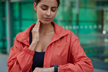 Cropped view of active sportswoman dressed in anorak checks pulse on neck after training looks at modern smartwatch poses against green blurred background. People workout and excersing concept