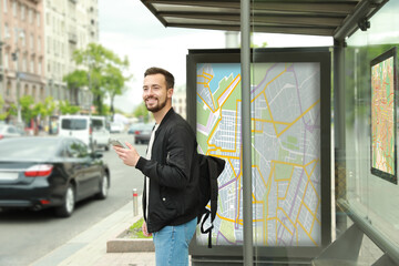 Young man with smartphone and backpack waiting for public transport at bus stop - obrazy, fototapety, plakaty