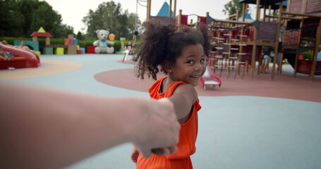Pov shot of little afro girl holding mother hand walking on playground - obrazy, fototapety, plakaty