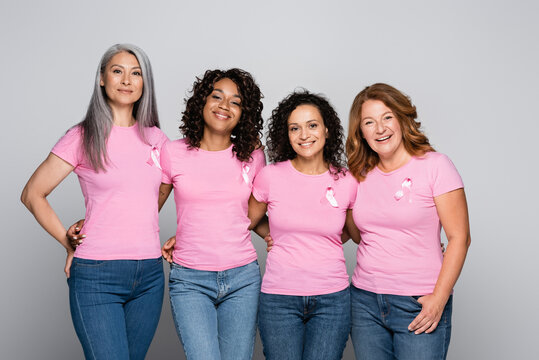 Multicultural women with ribbons of breast cancer awareness hugging on grey background