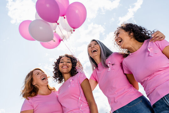 Low angle view of happy multiethnic women with ribbons of breast cancer awareness and balloons outdoors