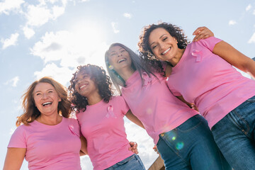 Low angle view of cheerful multicultural women with pink ribbons hugging outdoors
