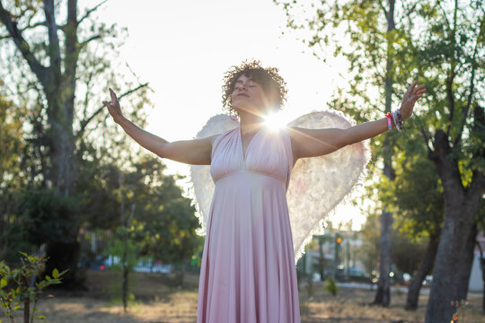 Mid adult afro mexican woman in a park standing with her eyes closed and her arms open, dressed in a pink dress and wearing wings like an angel
