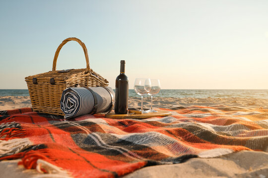 Blanket with picnic basket, bottle of wine and glasses on sandy beach near sea, space for text