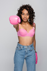 Fototapeta na wymiar Boxing glove on blurred african american woman in pink bra with ribbon isolated on grey