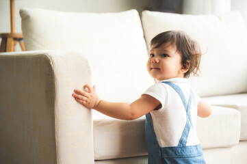 Cute little playful girl child learning to walk and stand by taking support of couch at home while looking away mischievously