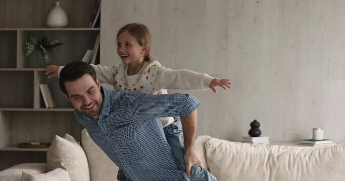 Happy father play with little daughter at home. Cute 9s girl hangs on daddy back enjoy active games in modern cozy living room, spread her arms imagining flying in air. Leisure, fun, travel concept