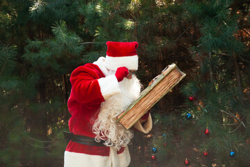 Santa Claus with a book in his hands against the background of Christmas trees studies it, reads...