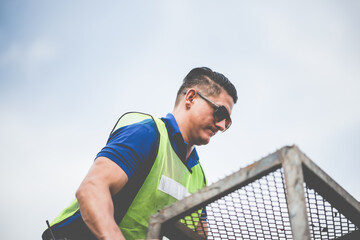 Fototapeta premium Young male warehouse supervisor wearing eyeglasses and vest supervising work holding hard hat in hand near waist