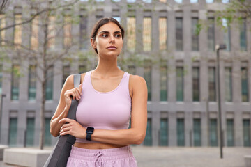 Thoughtful sporty woman in sportsclothes wears smartwatch for tracking fitness results carries karemat focused into distance poses outdoor against modern building. People sport workout concept