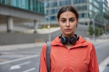 Horizontal shot of serious good looking sportswoman dressed in windbreaker uses wireless headphones returns from workout session after physical activities poses against blurred city background © WHstudio Leushin N