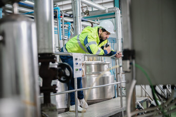 Young male machine inspector wearing vest and hardhat with headphones checking machine and sterilizers in water plant while making notes in digital tablet