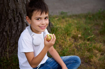 Adorable handsome schoolboy, elementary aged child sitting on the green grass of the park, eating an apple while resting during recreation after school and looking at the camera.