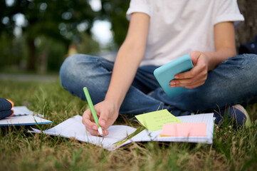 Cropped image of an unrecogniable school boy studying in the park, sitting on green grass and solving mathematics task, using smartphone and mobile applications, making notes on notebook and workbook.