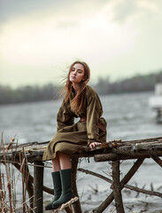 Lonely pensive girl, lost in thought, on wooden bridge by the river, in brown dress and rubber boots
