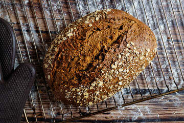 A loaf of whole wheat bread with cereal flakes, on an oven grid. With a oven glove.