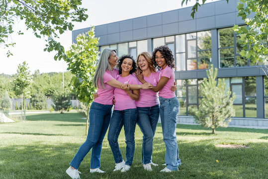 Cheerful multicultural women in pink t-shirts with ribbons hugging on grass