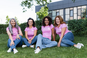 Happy interracial women with pink ribbons on t-shirts sitting on grass