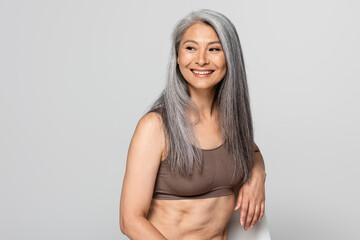 Positive asian woman in bra looking away on chair isolated on grey