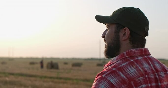 A bearded man in a baseball cap looks at a field after harvesting at sunset, a farmer inspects his field.