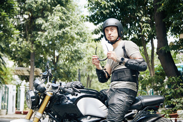Fototapeta na wymiar Serious man in helmet sitting on motorcycle and putting on glasses for protection