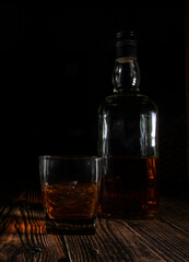 A glass of whiskey with ice and a bottle on a wooden table on a black background