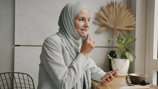 Happy hijabi girl is speaking on mobile phone wearing headphones holding smartphone in modern cafe. Cellphone and communication devices concept.