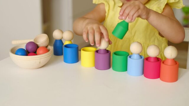 A two-year-old child plays with a wooden multi-colored toy sorter, studying colors. The girl takes out little man-shaped cylinders from the cups. Assemble the game according to the color