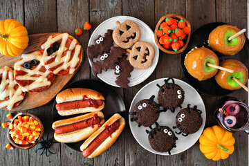 Halloween party food table scene over a rustic wood background. Overhead view. Spooky mummy pizzas, finger hot dogs, caramel apples, cupcakes, candy, cookies and donuts.
