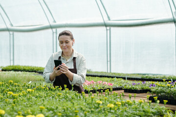 Happy female farmer with smartphone watching online gardening video in front of green seedlings