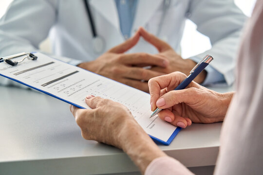 Close Up Shot Of Older Senior Female Patient Hands Holding Pen Filling Medical Heath Data Form Sitting At Desk With Family Doctor In Modern Hospital Clinic. Healthcare Illness Prevention Concept.