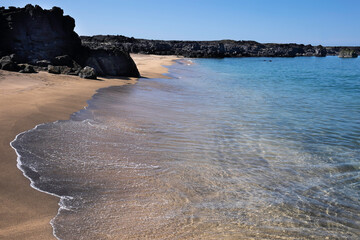 Empty sandy beach with crystal clear water and black lava stones and rocks on the coast of Iceland. Sunny day with blue sky
